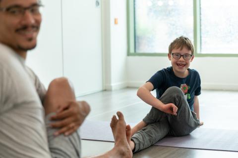 Young boy taking part in adaptive yoga, smiling to the camera. He is sitting on the floor with one leg straight out in front of him, the other leg bent and crossing over the straight leg with his hand holding the knee.