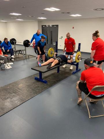 Stacey Denyer - Pictured lying on a weights bench in an indoor gym environment, holding a laden bar bell, resting across her chest. She has 3 people spotting, 1 behind her head and 1 either side of the bar bell. Other People are stood around watching.