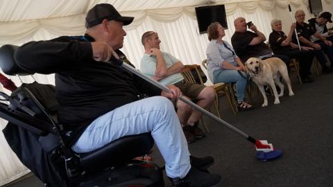 Wheelchair user taking part in a floor curling activity at Dragons RFC 'Sporting Memories' session. He is sat in his wheelchair on the left of photo facing to the right. He is holding a curling pole with the curling stone on the floor ready to be pushed.