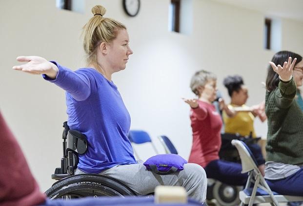 Wheelchair user with blond hair tied up in a top knot wearing a blue shirt extending her arms out to the side with a sandbag on her lap to help ground her in the yoga pose , other students, one in a red top, one in a yellow top an one in a green top all mimic the pose in the background 